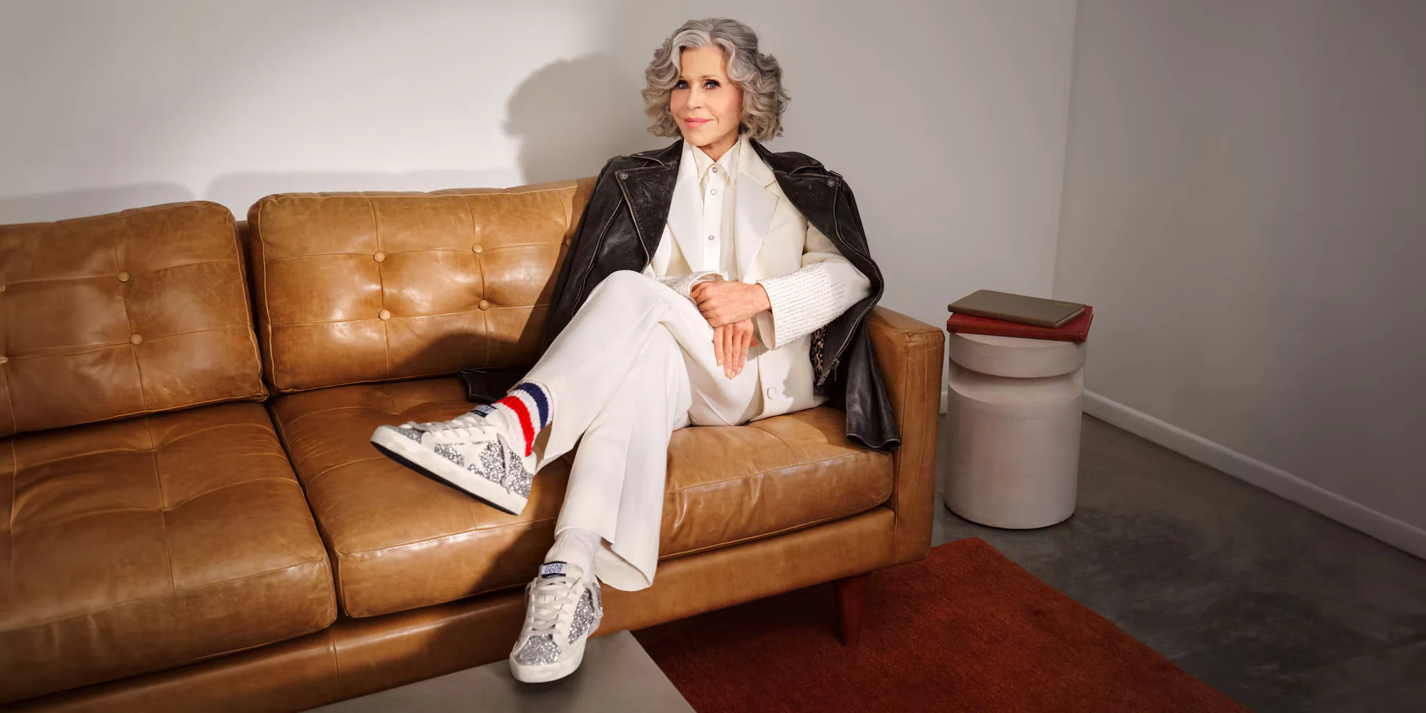Jane Fonda wearing Golden Goose Super-Star sneakers on a tan sofa.
