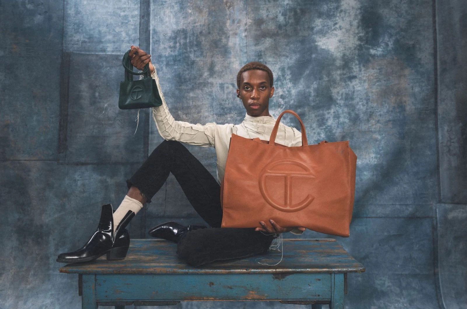 Model displaying black and tan Telfar Shopping Bags on a blue table.