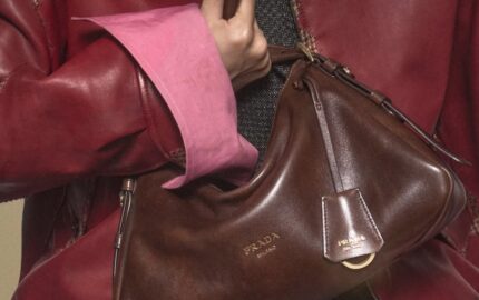 Close-up of a brown antiqued leather Prada shoulder bag with embossed logo, worn under the arm with a red leather jacket