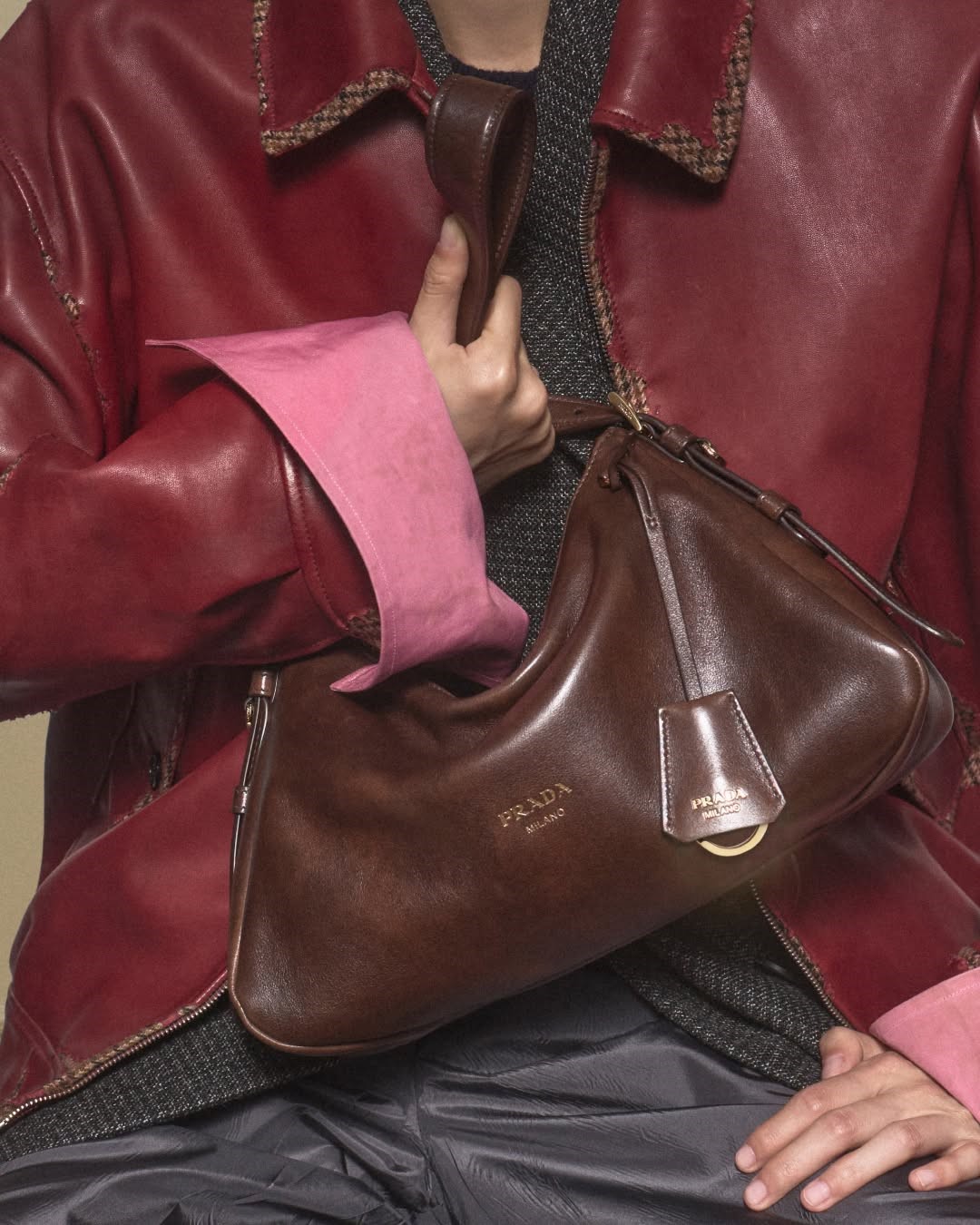 Close-up of a brown antiqued leather Prada shoulder bag with embossed logo, worn under the arm with a red leather jacket