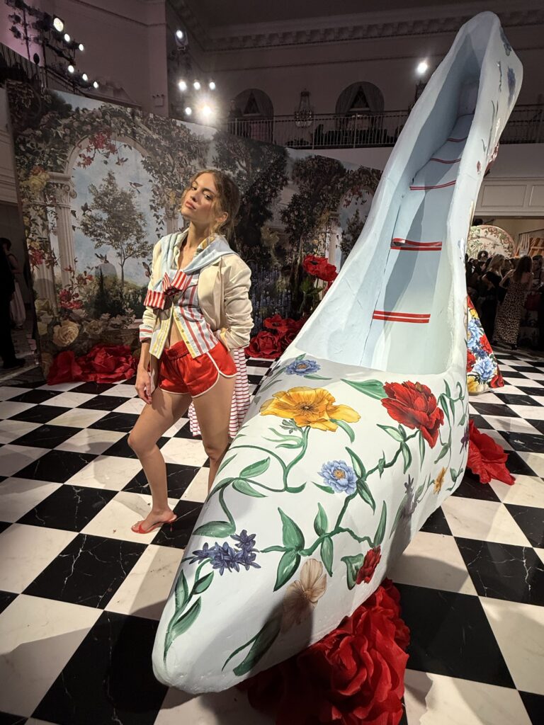 Model wearing a layered striped look beside an oversized floral shoe sculpture at Alice + Olivia Spring/Summer 2027.