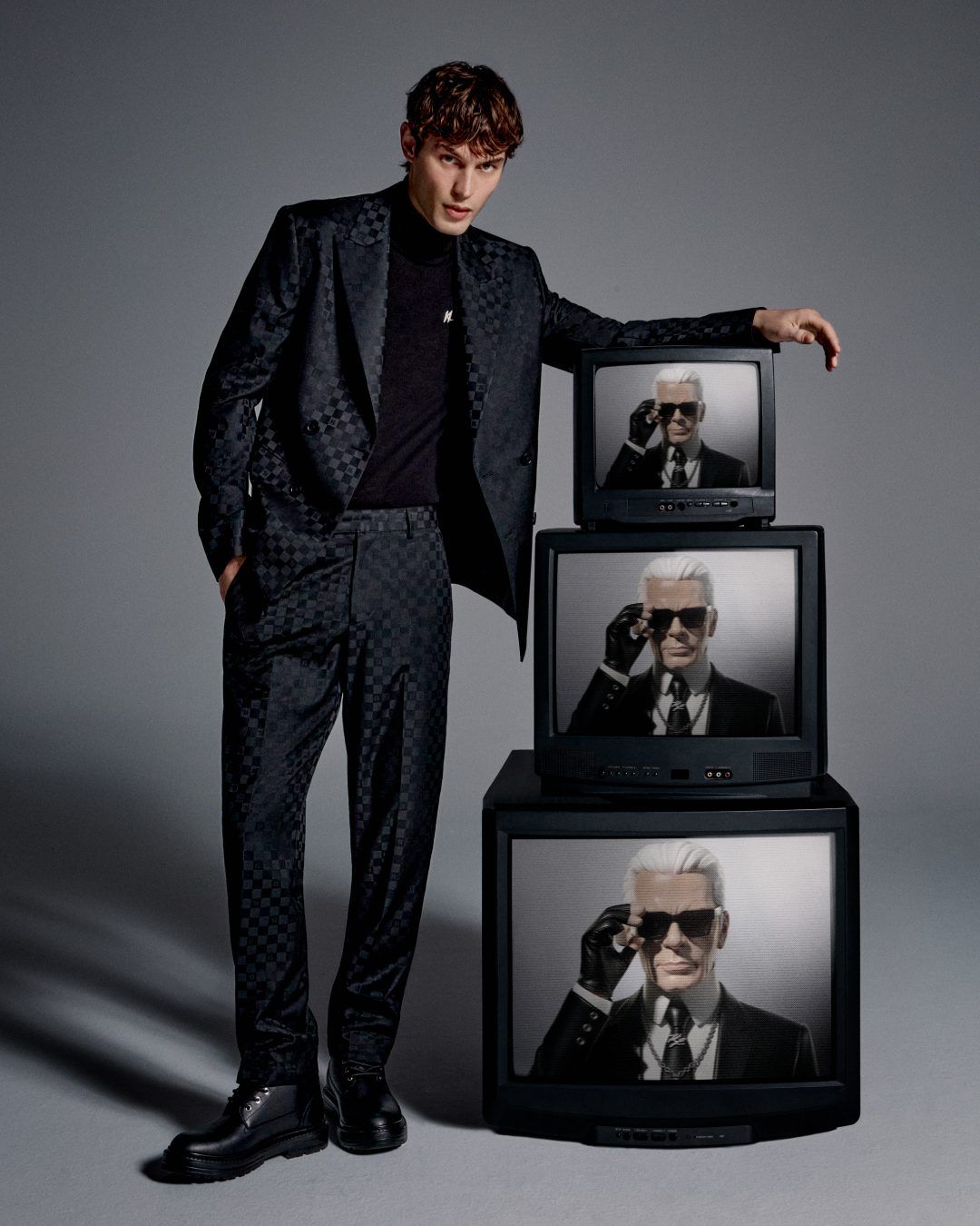 Kit Butler wearing patterned black tailoring beside stacked television screens