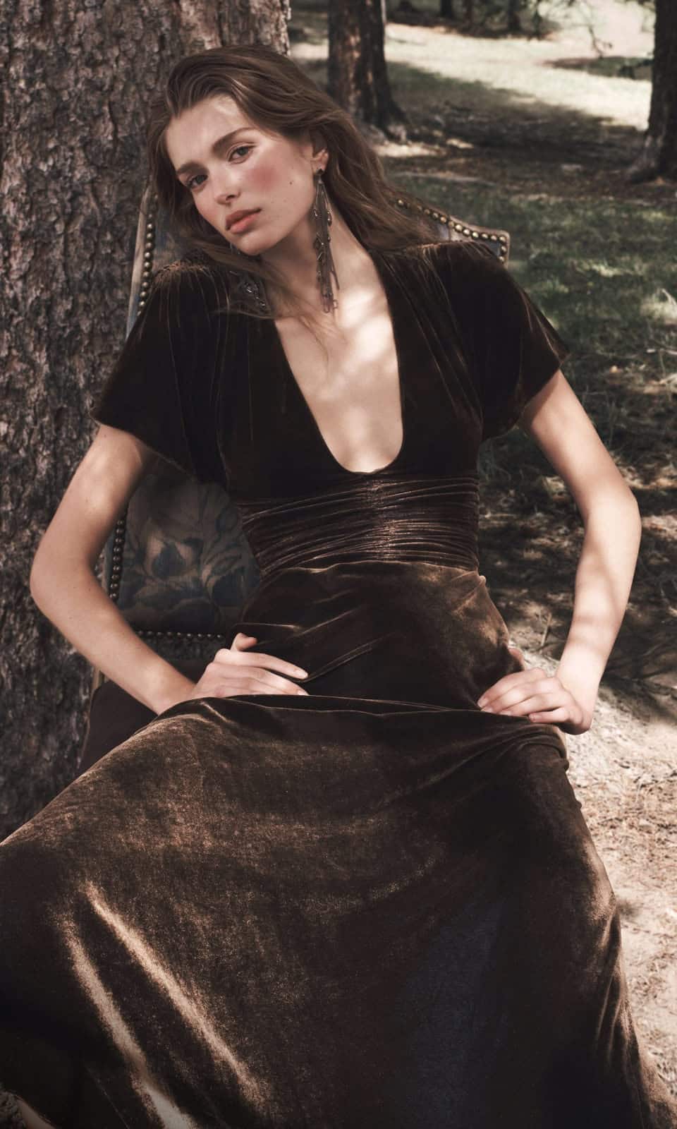 Model in a dark velvet dress seated outdoors beside a tree.