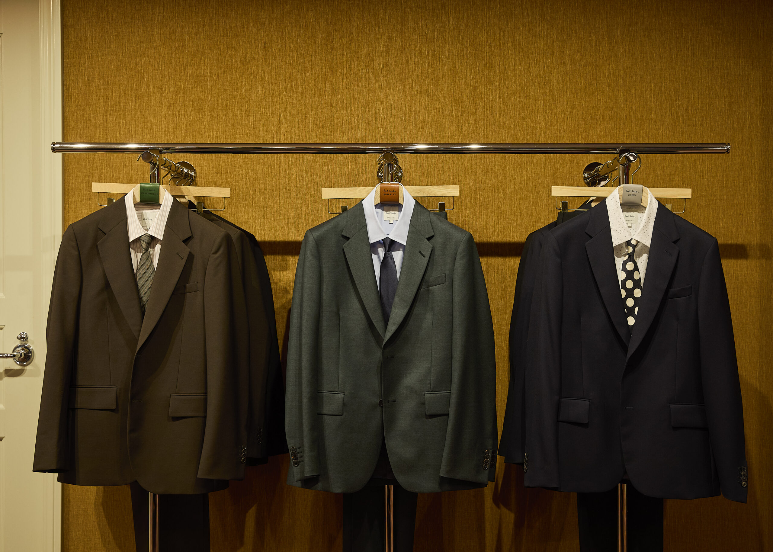Three tailored suits displayed against a textured wall at Paul Smith Madison Avenue