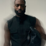 Dwyane Wade wears a black sleeveless shirt and holds a black woven bag in a studio portrait.