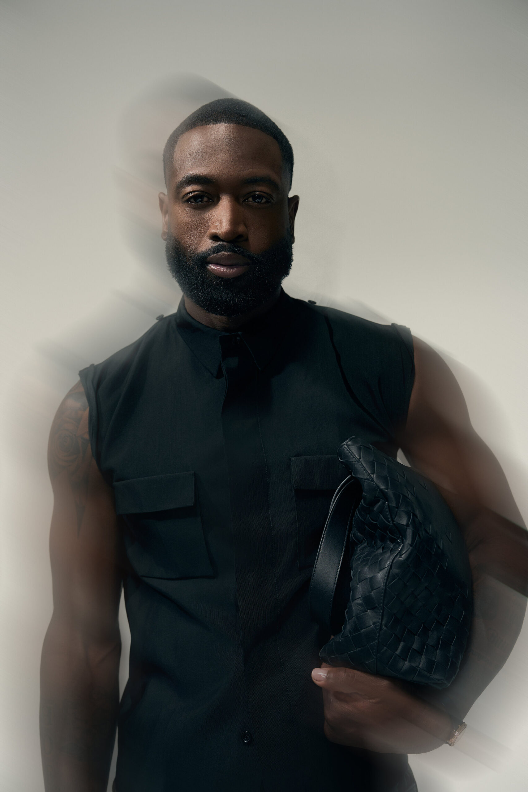 Dwyane Wade wears a black sleeveless shirt and holds a black woven bag in a studio portrait.