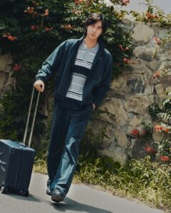 Wang Yibo in a blue jacket and striped polo pulling a suitcase beside a stone wall