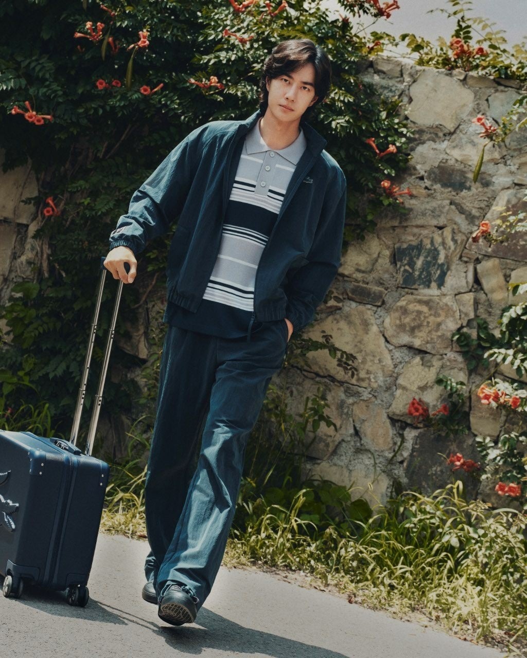 Wang Yibo in a blue jacket and striped polo pulling a suitcase beside a stone wall