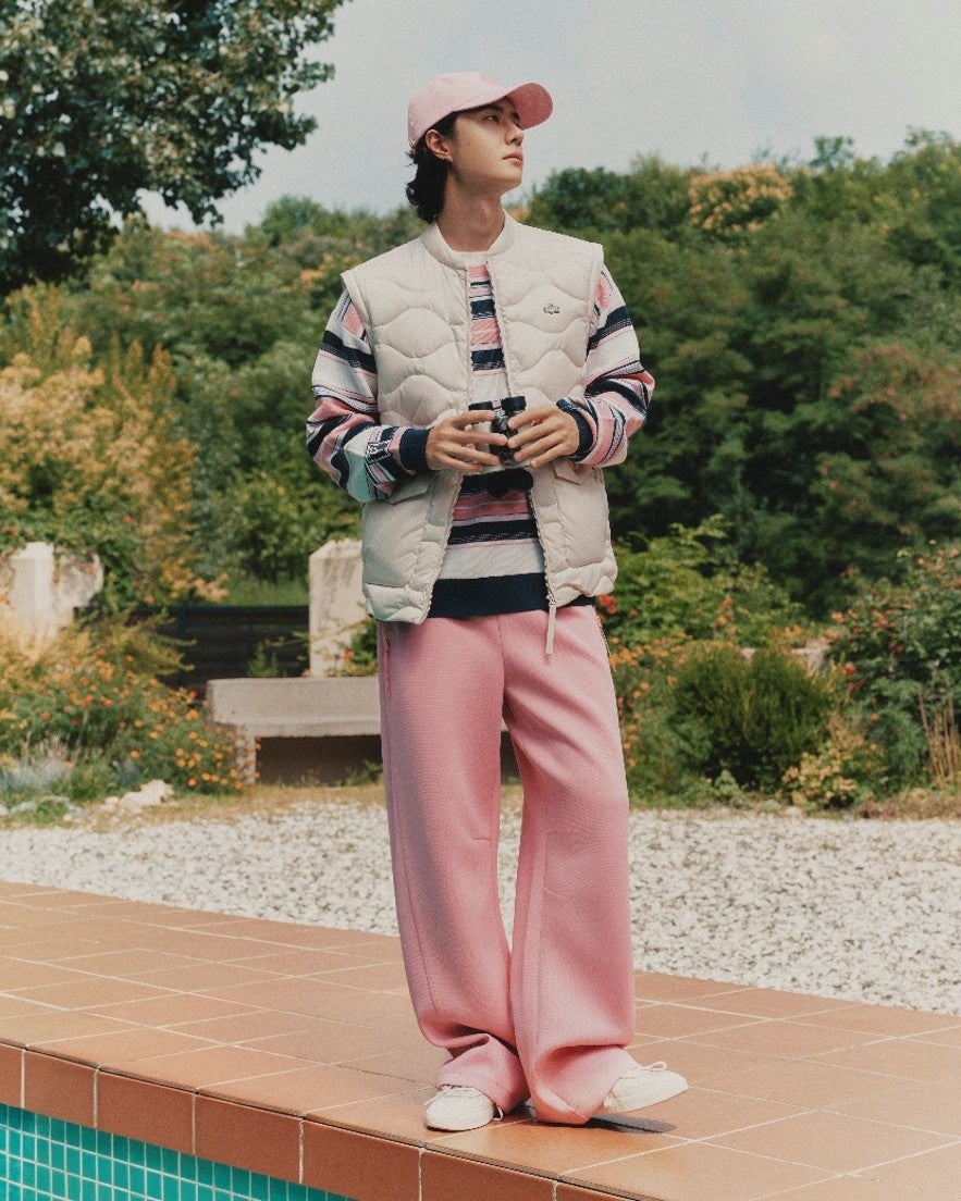 Wang Yibo wearing a cream quilted vest, striped knit and pink trousers beside a pool