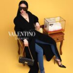 Jolin in black Valentino clothing beside a display case against a yellow backdrop