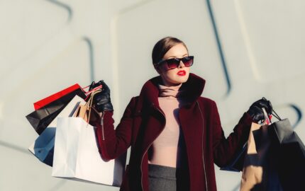 Woman in sunglasses and a burgundy coat holding several shopping bags