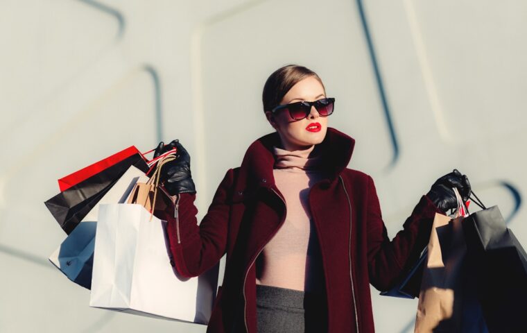 Woman in sunglasses and a burgundy coat holding several shopping bags