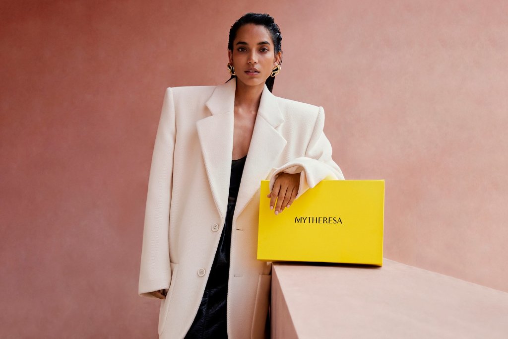 Model in a white suit holding a yellow Mytheresa shopping bag