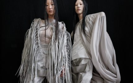 Two fashion models in sculptural white outfits against a black backdrop