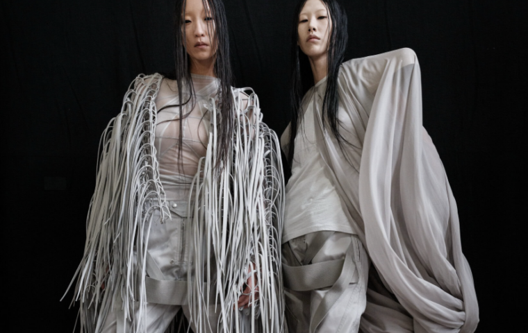 Two fashion models in sculptural white outfits against a black backdrop