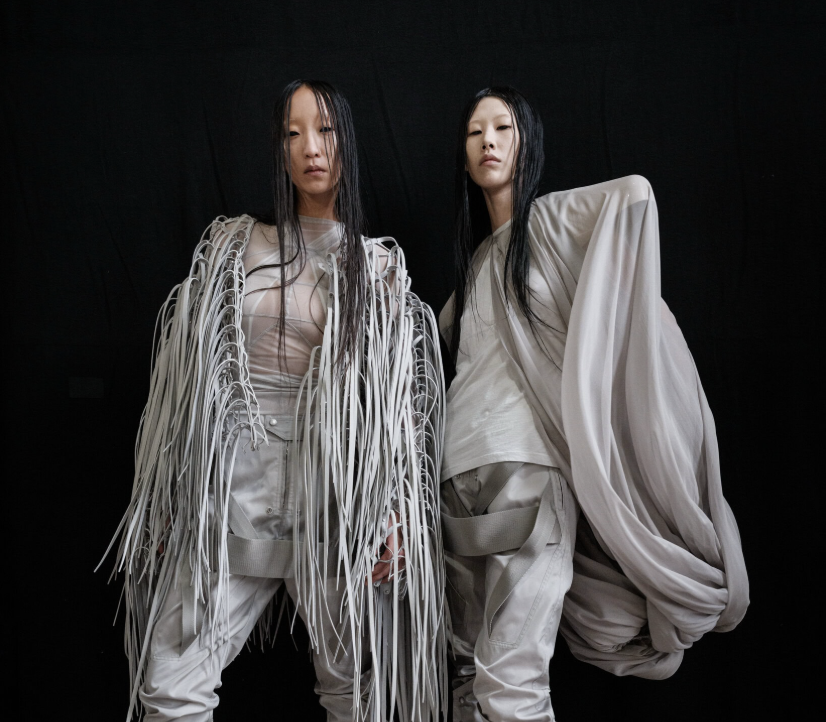 Two fashion models in sculptural white outfits against a black backdrop